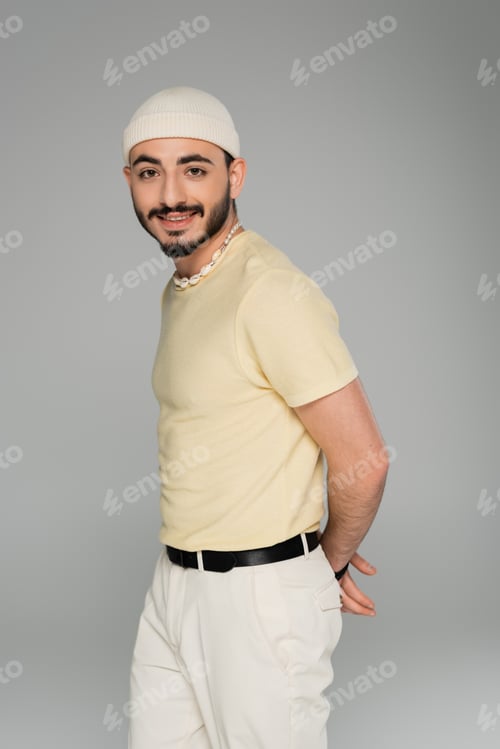 Preview: Portrait of trendy homosexual man in hat and seashell necklace smiling at camera isolated on grey