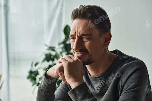 Preview: upset bearded man in casual home wear feeling unwell and frowning while looking away at home