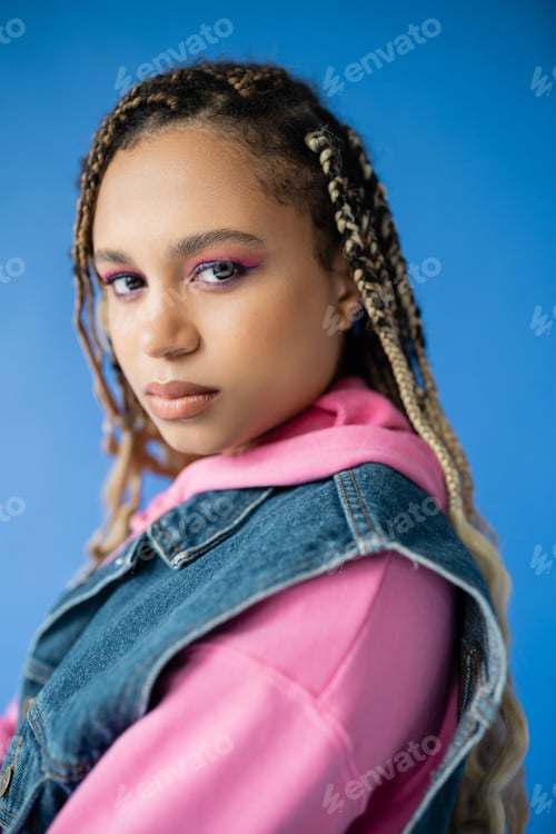 Preview: attractive african american woman with dreadlocks looking at camera on blue background, bold makeup
