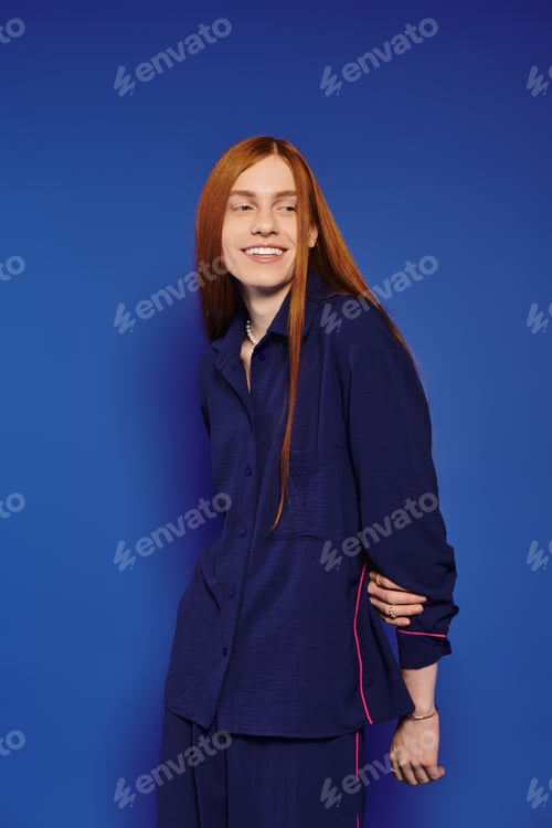 Preview: Handsome young queer man with long red hair poses stylishly against a vibrant blue backdrop