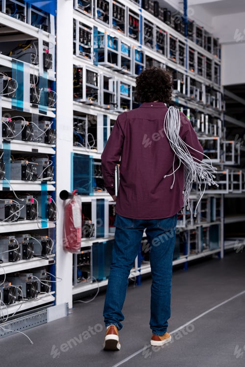 Preview: rear view of computer engineer with ethernet wires on shoulder at cryptocurrency mining farm