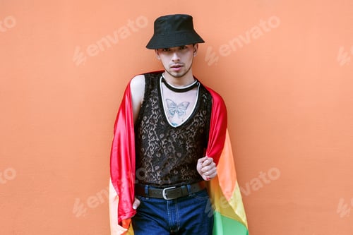 Preview: Handsome young man in lgbt community hat draped with gay flag on a coral colored wall.