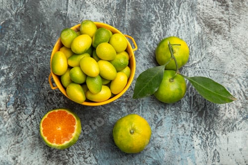 Preview: Overhead view of a yellow bucket full of fresh green tangerines and cut in half tangerines on gray