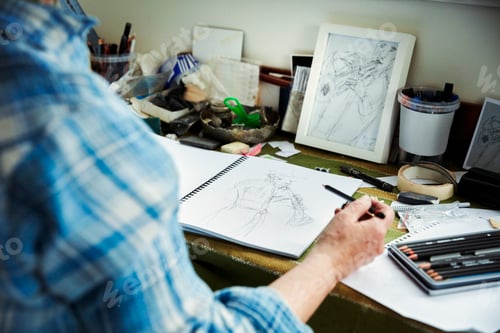 Preview: An artist working, holding a pencil over a sketch in progress in an open sketchbook.