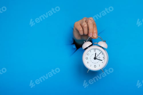 Preview: female hand holds pink alarm clock through a paper hole in blue background.