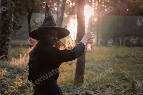 Preview: Young woman in dark dress and witch's hat holds lantern with candles in hands and illuminates path