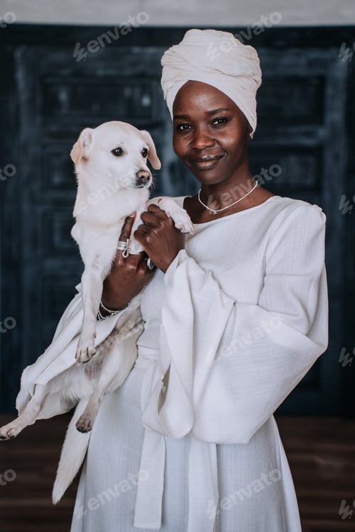 Preview: Vertical shoot of African young woman in white turban and traditional white dress holding dog home