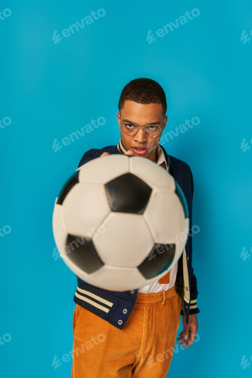 Preview: confident and serious african american student showing soccer ball at camera on blue