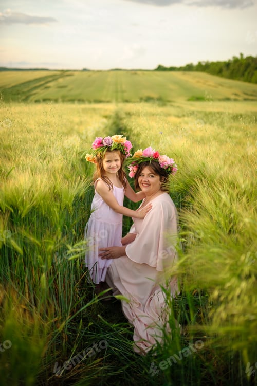 Preview: Mother and daughter in pink dresses in a green wheat field. On their heads they have floral wreaths.