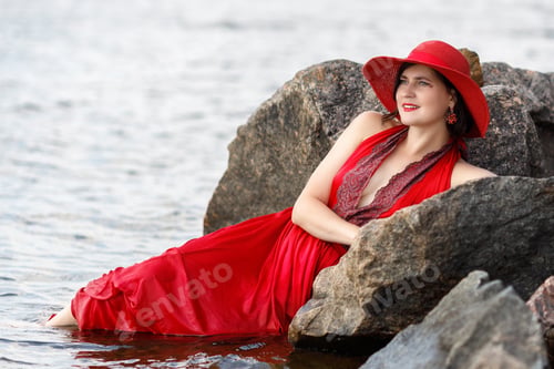 Preview: A beautiful elegant lady in a red hat and a long red dress is swimming in the water near the stones