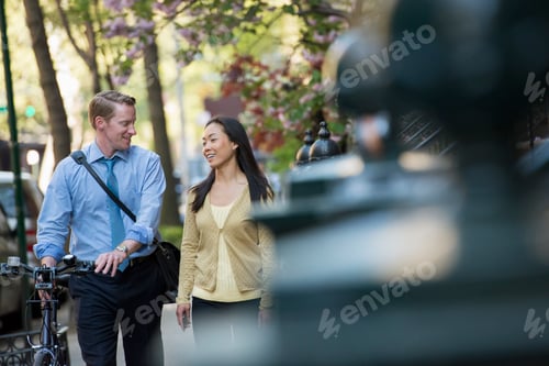 Preview: A man in a shirt and tie pushing a bicycle, and talking to a woman. A park in the city.