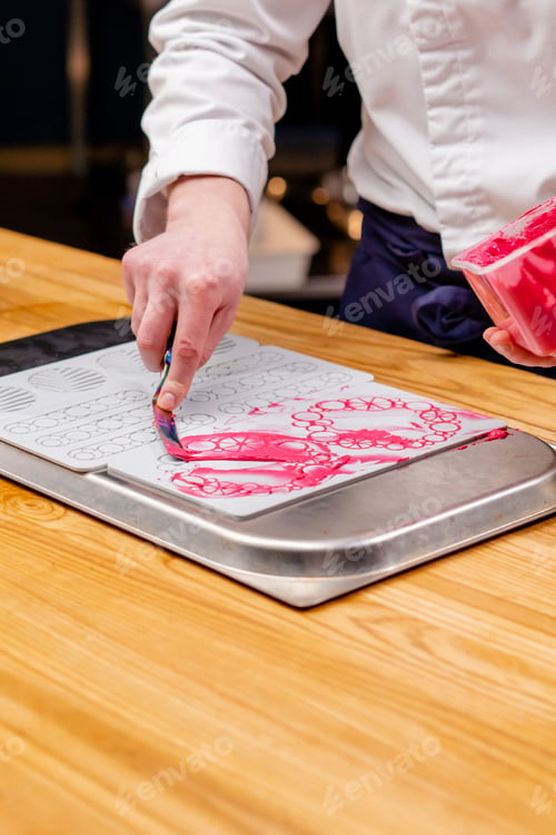 Preview: close-up of hands at the table spreading pink sweet spread onto mold with a spatula