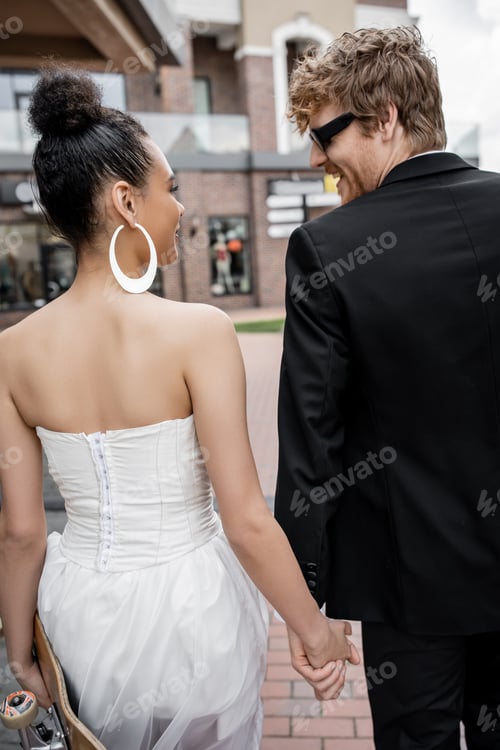 Preview: redhead groom in sunglasses, african american bride with longboard looking at each other on street