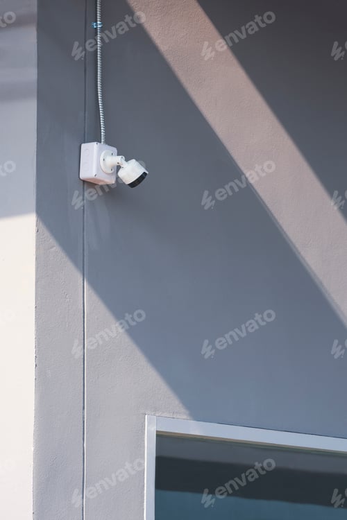 Preview: security camera and flexible metal conduit with part of glass window on gray cement wall