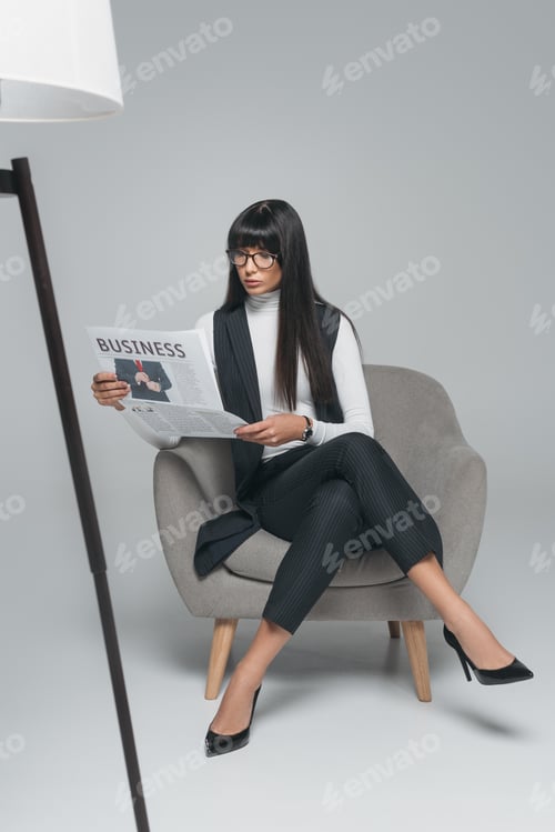 Preview: beautiful brunette businesswoman reading newspaper in armchair on gray