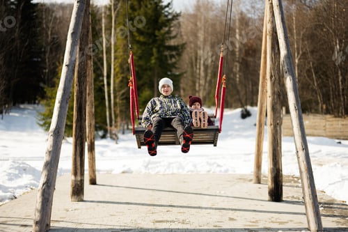 Preview: Older brother with younger sister swinging on big wooden swing in forest of mountains.