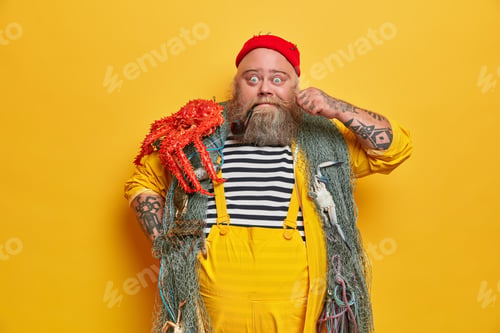 Preview: Indoor shot of bearded surprised man mariner curls mustache, smokes tobacco pipe and poses with fish