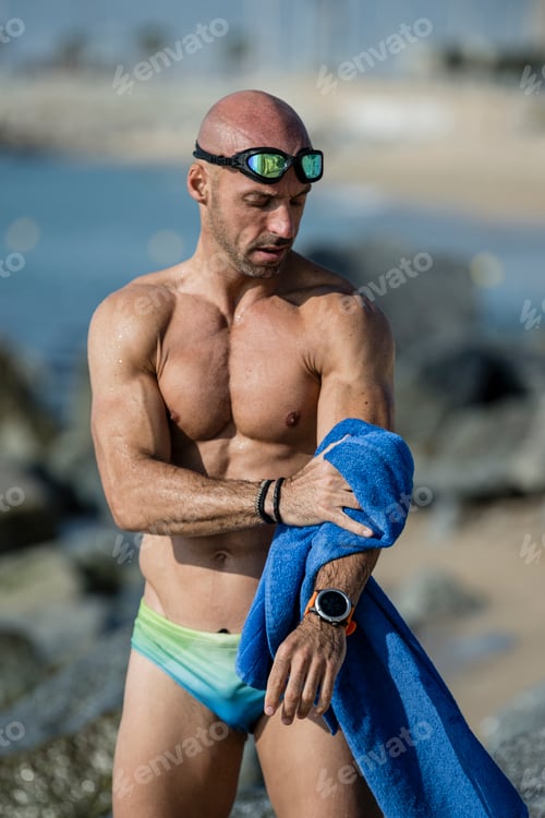 Preview: Muscular swimmer toweling off his arm with swim glasses at the beach. Sexy fit man with a blue towel