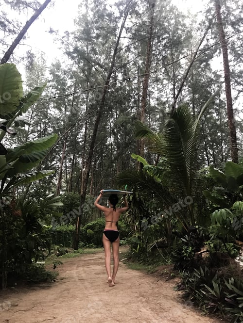 Preview: Woman Walking Barefoot with Surfboard in Tropical Forest