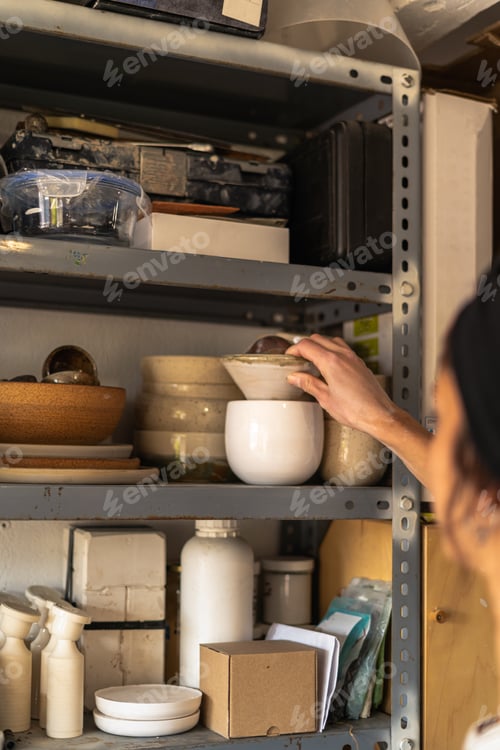 Preview: Young female potter arranging ceramic products on workshop shelf
