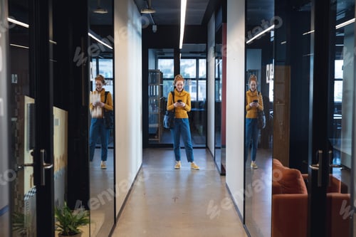 Preview: Mid adult caucasian businesswoman using smartphone while standing in corridor of creative office