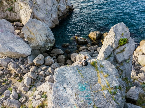 Preview: aerial directly above view of young man sitting on the mountain edge rock with backpack