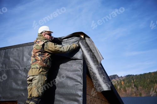 Preview: Male builder installing waterproof membrane on wall of future wooden frame house.