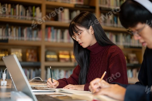 Preview: Glasses asian woman student writing in notebook beside a man friend while sitting at library's table