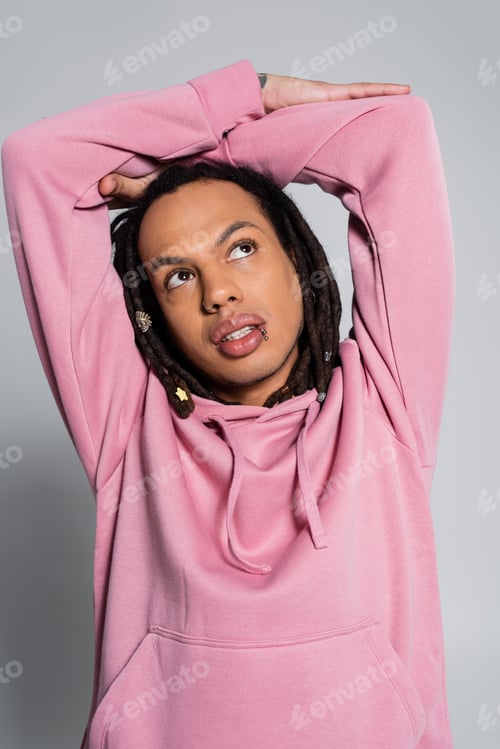 Preview: tattooed multiracial man in pink hoodie looking up while thinking on grey
