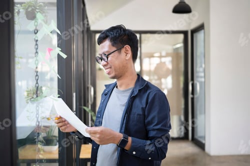 Preview: Smiling young creative designer reading document while standing near glass wall in modern office.