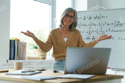 Preview: Cheerful senior woman gesturing while standing near the whiteboard at classroom