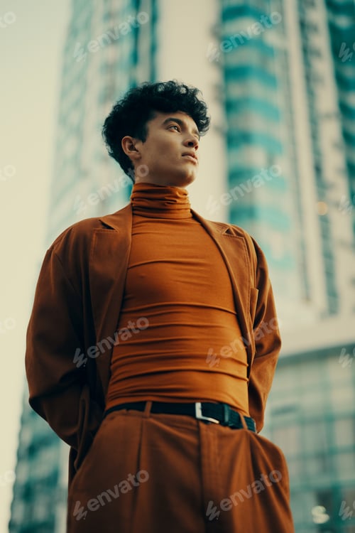 Preview: Young man in stylish brown outfit stands confidently against modern skyscrapers in a vibrant urban
