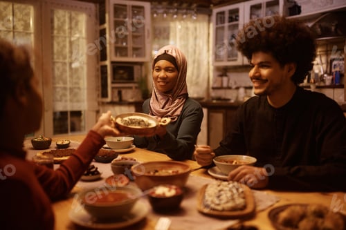 Preview: Happy Muslim woman passing food to her daughter during family dinner at dining table.