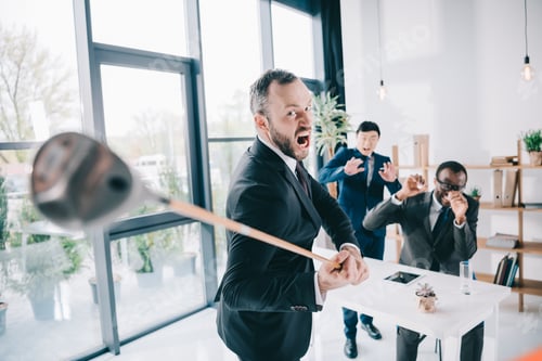 Preview: angry businessman with golf cub looking at camera with his scared colleagues on background