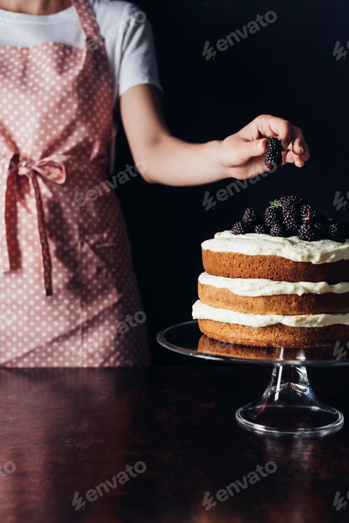 Preview: cropped shot of woman decorating freshly baked blackberry cake on glass stand on black
