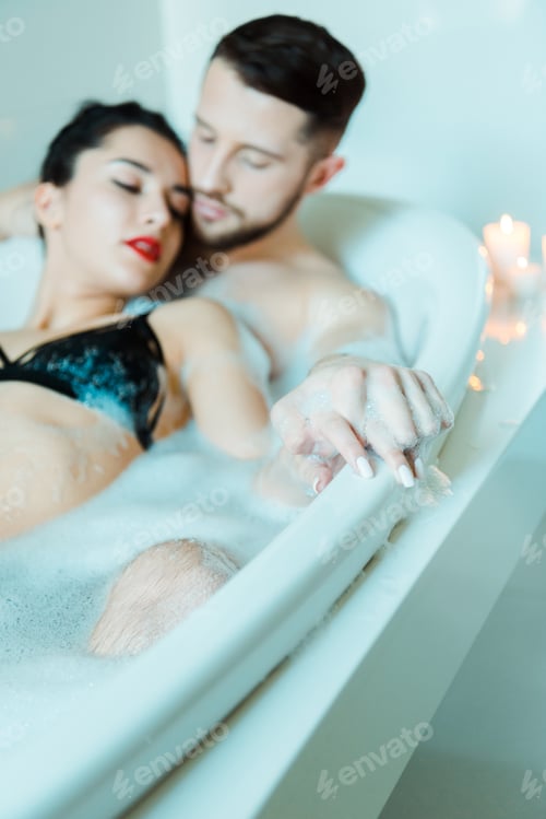 Preview: selective focus of bearded man and attractive brunette woman holding hands while lying in bathtub
