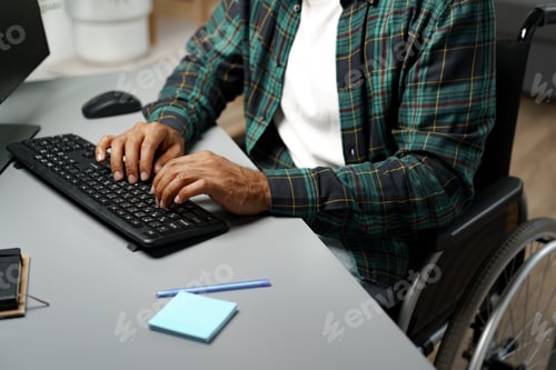 Preview: Disabled young african american man in wheelchair using computer while sitting at his working table