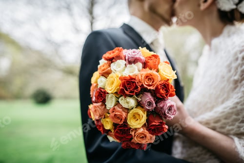 Preview: Closeup shot of the bride's bouquet with colorful flowers and the couple kissing in the back