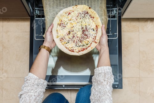 Preview: Woman placing a ham and cheese pizza into an oven from a bird's-eye perspective in a modern kitchen