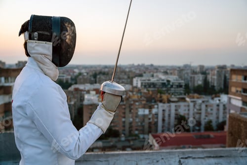 Preview: Portrait of young woman fencer wearing mask, white fencing costume and practicing outdoors.
