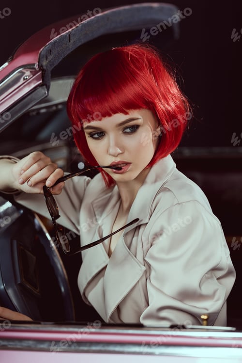 Preview: beautiful young woman in red wig holding sunglasses and looking away while sitting in car