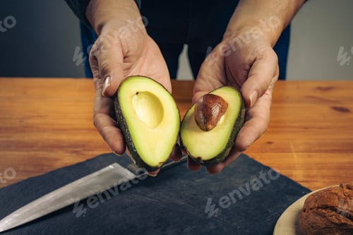 Preview: Male hands holding a fresh avocado cut in half with pit, over a wooden table.