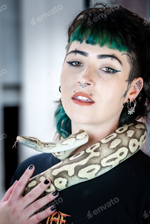 Preview: Alternative girl with coloured hair and facial piercings posing with a Ball Python snake