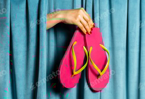 Preview: Female hand holds pink and green flip flops on a background of blue velvet curtains
