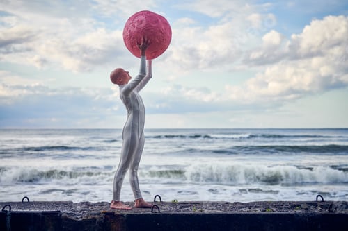 Preview: Hairless girl with alopecia in white futuristic suit standing on concrete fence on sea background