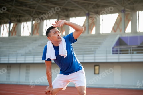 Preview: Asian man in blue t shirt is standing with hands on his knee and looking up with hand over eyes.