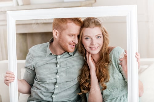 Preview: Beautiful young couple sitting embracing on sofa and looking through white frame