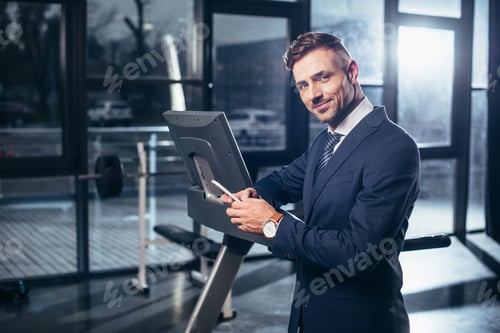 Preview: handsome businessman in suit leaning on treadmill and using smartphone in gym