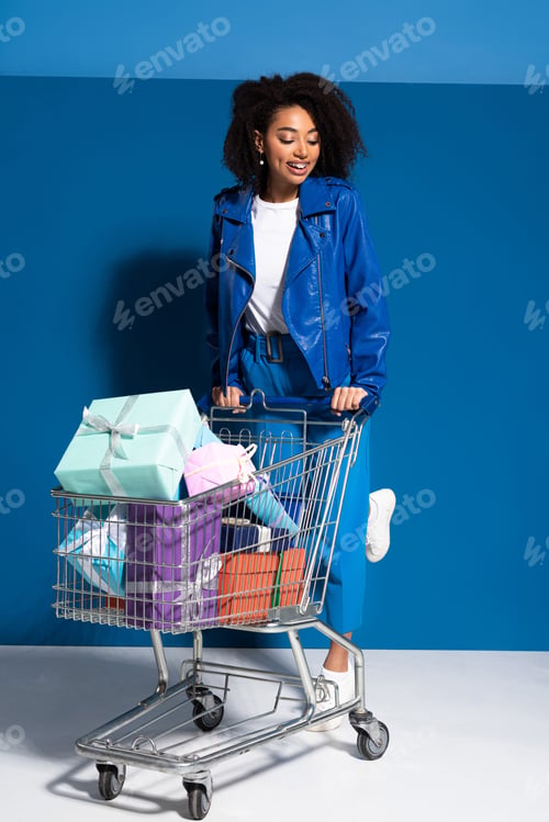Preview: smiling african american woman with shopping cart full of gifts on blue background