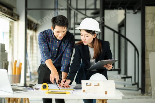 Preview: Two young Asian architects discussing building plans with laptop, scale model, and hardhat